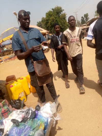 The image depicts an outdoor market scene in Kpak, Nigeria, under clear daylight. In the left foreground, a dark-complexioned male with a shaved head and black sunglasses pushed onto his head wears a blue short-sleeved collared shirt, dark pants, and a brown leather shoulder bag. He is holding and examining a stack of currency notes with both hands. To his left, three yellow plastic jerrycans, at least one branded "OKD" with an image of a fowl, are visible on the ground. In the immediate foreground, a vendor's cart or display stand is filled with assorted items packaged in clear and colored plastic bags.
To the right of the primary male subject, two other dark-complexioned males are walking. The one closer to the center wears a light-colored t-shirt with brown and black stripes and "Happy New Year" text. The one behind him wears a black kaftan with white embroidery. A fourth male, partially visible on the far right, wears a white t-shirt with blue striped sleeves. The ground is dry, unpaved light brown earth. In the background, market stalls with produce, including red items, are visible under a blue and yellow umbrella. Simple structures with corrugated metal roofs, along with utility poles and wires, line the distant background.