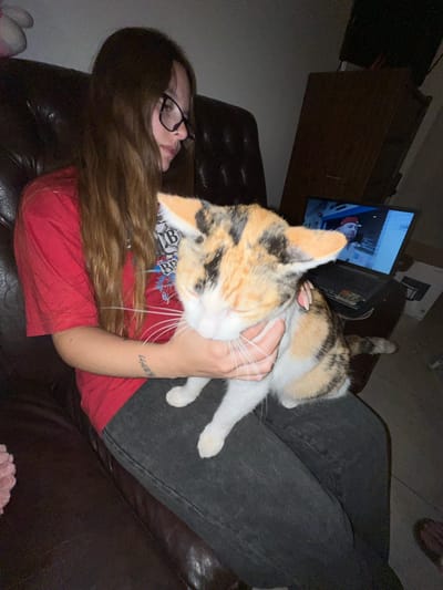 A woman with long brown hair and glasses is lying on a dark brown leather couch, wearing a red shirt and dark jeans. She is holding a calico cat with her right hand, its head resting on her chest. The cat has a mostly white face with patches of orange and black fur. In the background, there is a dark brown wooden cabinet and a television displaying an image of a person wearing a red and white hat. The location is unknown and cannot be determined to be in Bahía Blanca, Argentina based on the provided visual information.
