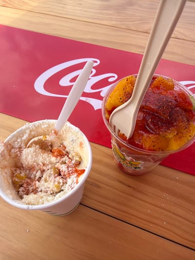 Two cups of what appear to be sweet and savory snacks are placed on a wooden table. To the right, a clear plastic cup is filled with sliced fruit, possibly mango, topped with a red sauce and sprinkled with chili powder. A beige plastic spork is inserted into this cup. To the left, a white paper cup holds a creamy mixture, topped with shredded white cheese and red seasoning. A white plastic spoon is in this cup. A red placemat with a white Coca-Cola logo is partially visible beneath the cups. The lighting suggests an outdoor setting. The image was taken in Houston, Texas, United States of America.