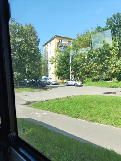 The image depicts a scene in Zhukovsky, Russia, likely in a residential area, as indicated by the presence of a multi-story apartment building and parked cars. The apartment building is yellow and appears to have balconies. Lush green trees and grass surround the building and the paved road. Several vehicles are visible: a dark sedan, a white pickup truck, and a white car with colorful decals, possibly a service vehicle. There is a painted bicycle symbol on the paved pathway, suggesting a designated cycling route. The sky is clear and blue, indicating fair weather. The image is taken from the interior of a vehicle, as suggested by the dark frame on the left side and the reflection on the windows.