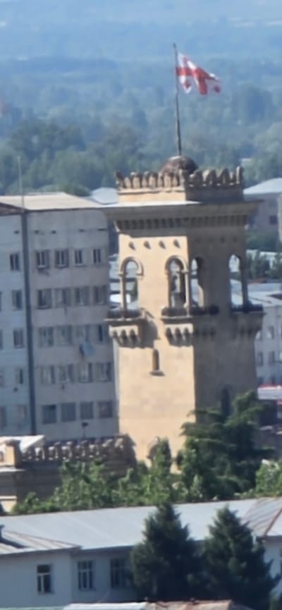 The image shows a view of a building with a flag on top, located in Gori, Georgia. The building appears to be a historical or significant structure, with crenellations and arched windows. A flag, likely the flag of Georgia, is flying from a flagpole on the building. In the foreground, there is a tall, modern building with many windows. The background shows a landscape of trees and hills, suggesting an outdoor setting on a clear day. The time of day appears to be daytime, likely morning or afternoon, based on the lighting. There is no visible text in the image. The overall impression is one of a cityscape with a prominent landmark.