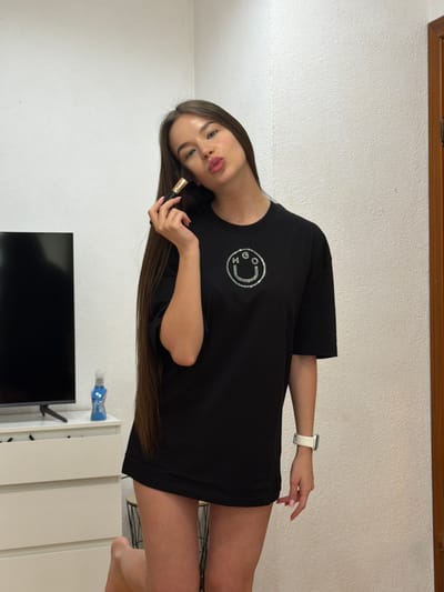 A woman is posing in a room. She is wearing a black t-shirt with a smiley face design. She is holding a makeup brush in her hand. The room contains a television, white cabinets, and a wooden trim. The city of Kovel, Ukraine, is not identifiable from the image.