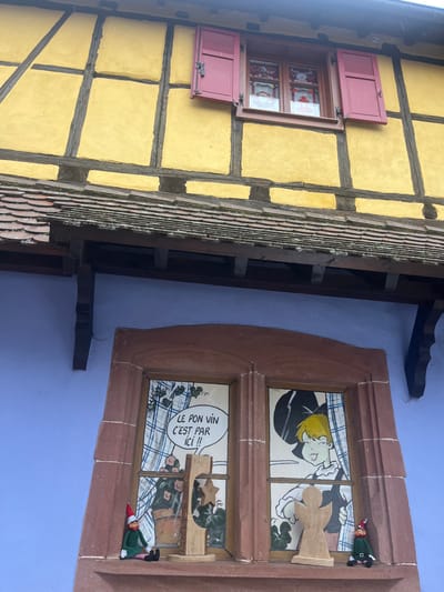 A two-story building facade in Kintzheim, France, is depicted from a low-angle perspective. The upper story exhibits a half-timbered construction with yellow infill panels and dark brown structural beams. A single rectangular window on this upper level features dark red wooden shutters; the left shutter is closed, while the right shutter is open, revealing a dark-framed window with white curtains patterned with red designs. Below the upper story, a terracotta-tiled roof with dark wooden supporting corbels extends over the lower section of the building. The lower story is painted a solid periwinkle blue. Centered on this blue wall is a large, rectangular window with a prominent reddish-brown stone frame. This window is divided into two panes. The left pane displays a comic-style illustration featuring a speech bubble with the French text "LE BON VIN C'EST PAR ICI !!" (The good wine is here!!) above a depiction of a potted plant with pink flowers. The right pane shows a comic-style illustration of a person with blonde hair and a dark hat, appearing to smile or wink. On the exterior sill of this lower window, two small figures resembling elves or gnomes, dressed in green and red, are seated. Between them are two wooden decorative cutouts: a stylized wooden Christmas tree with a yellow star on the left, and an unpainted wooden angel cutout on the right.