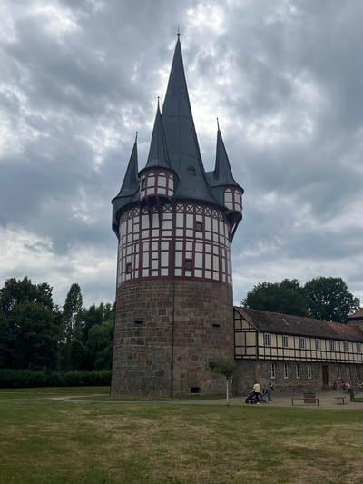 This image captures a view of a historical tower with a distinctive half-timbered upper section and a stone base. The tower features multiple conical roofs, the tallest of which is a dark gray slate. The half-timbered section is white with dark red beams, forming a crisscross pattern.

To the right of the tower, a building with similar half-timbered architecture is visible. This building has a red-tiled roof and multiple windows. The area around the tower and building is a grassy lawn.

In front of the building, several people are seen. Two individuals are pushing strollers, and a few others are seated on a bench or in the background. The scene suggests a daytime visit to a historical site or park. The sky is overcast with gray clouds, indicating cloudy weather.

The location is Neustadt, Germany, which is suggested by the architectural style commonly found in this region. The overall atmosphere is peaceful and leisurely, with visitors enjoying the grounds. There is no visible text in the image.