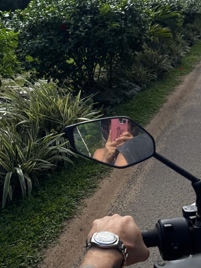 In Olaganduwa, Sri Lanka, a person is riding a motorcycle on a paved road flanked by lush vegetation. The rider, reflected in the motorcycle's rearview mirror, is holding a pink smartphone with a black case and is wearing a helmet. The mirror also shows a portion of the rider's arm and shoulder, and a glimpse of foliage in the background. The rider's left hand, wearing a silver watch, is on the motorcycle's handlebar. The environment is verdant, with dense green bushes and trees lining the road.