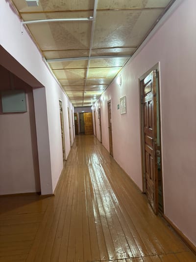 This image depicts a long, unoccupied indoor hallway within a building in the city of Bayronovka, Russia. The hallway extends into the distance, framed by light pink walls on both sides. The floor is composed of varnished, light brown wooden floorboards, reflecting light from overhead fixtures. The ceiling features a grid of white metal framing, containing rectangular panels of a light-colored material with a subtle pattern, possibly wood grain or a decorative texture.
Multiple identical wooden doors, each with recessed panels, line both walls of the hallway, receding in perspective. Most doors appear closed, but one door on the right side in the foreground is slightly ajar, revealing a dark interior beyond. Wall-mounted light fixtures with warm illumination are spaced along both walls. On the left wall, near the foreground, a white rectangular electrical panel cover is visible. Further down the hallway on the right wall, a white sign with a green symbol and Russian text, "ВЫХОД" (EXIT), is mounted. No individuals are present, and no actions or interactions are depicted. The scene is illuminated by artificial light, casting reflections on the glossy floor surface.