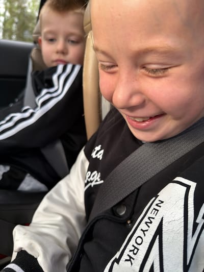 The image shows two children in a car. One child, with closed eyes and fair skin, is wearing a black and white "New York" jacket and a seatbelt. Below, another child is visible in a black Adidas tracksuit. The location appears to be within a vehicle, with exterior elements possibly visible outside the window. Specific details about the city of Savinskoye Sel'skoye Poseleniye, Russia are not discernible.