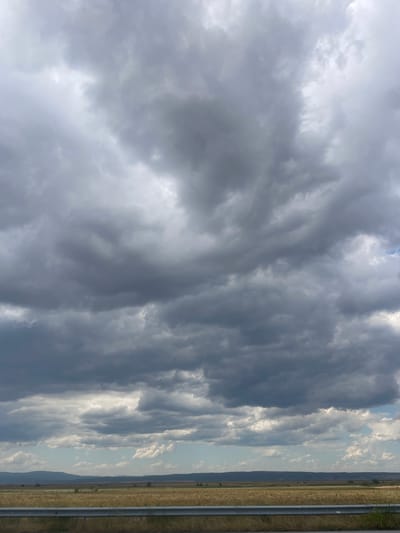This is a photograph taken from the side of a road, looking out over a flat, rural landscape. The sky dominates the frame, filled with dramatic, dark, and heavy storm clouds. The clouds are textured and appear to be moving, with varying shades of grey and some areas of lighter grey where the sun is attempting to break through.

Below the clouds, the horizon is defined by a low, rolling line of hills or mountains in the distance. In the middle ground, there is a wide expanse of dry, golden-brown fields, possibly crops like wheat or barley, stretching towards the hills. Scattered trees are sparsely visible within the fields.

In the foreground, a metallic guardrail runs horizontally across the bottom of the image, indicating the presence of a road. Beyond the guardrail, there is a strip of dry grass and vegetation. The overall impression is one of an approaching storm in a rural, agricultural area. The location is stated to be in the city of Zheleznik, Bulgaria. There are no people or animals visible in the image.