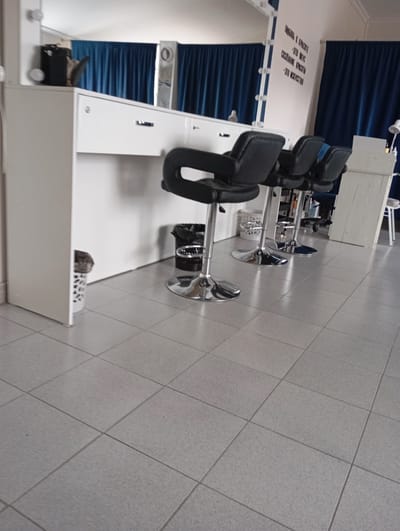 The image depicts a makeup studio in Nazran, Russia. The scene features a row of three black leather salon chairs with chrome bases positioned in front of a white counter. Behind the chairs, a white vanity with mirrors is visible, framed by blue curtains. A black trash can with a chrome rim sits to the left of the chairs. On the counter, text is written in Russian, which translates to "Love for beauty is taste. Creation of beauty is art." A white waste bin with a patterned design is situated to the left of the counter. The floor is covered with light gray square tiles.