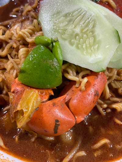 A close-up, high-angle shot depicts a bowl of prepared food, likely Mie Aceh, situated in Alue Mudem, Indonesia. The central subject is a portion of reddish-brown broth containing numerous curly noodles. Prominently placed in the foreground is a bright orange crab claw or shell segment. Garnishing the dish are two thin, translucent slices of cucumber positioned at the top right, a wedge of bright green lime directly above the crab, and two small, green chili peppers adjacent to the lime. The broth exhibits a somewhat oily texture and contains fine particulate matter. A white bowl rim is visible in the bottom left corner, indicating the container. No people or specific environmental details beyond the dish itself are discernible.