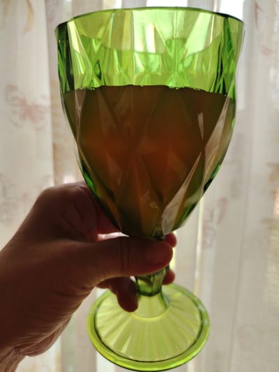 A hand is holding a green glass goblet filled with a dark amber liquid. The goblet has a faceted design, and the liquid appears to have some small bubbles at the surface. The background consists of a sheer, light-colored curtain with a subtle floral pattern, suggesting an indoor setting, possibly near a window. The lighting indicates daylight. The location of Кривец, Russia is not evident from the image content.