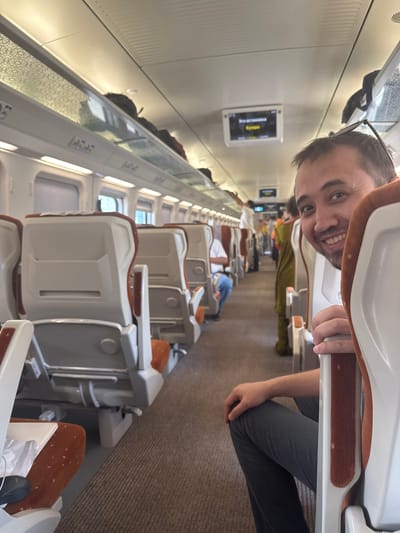 The image shows the interior of a modern train carriage, likely in Uzbekistan. The primary subject is a man with a beard and glasses, who is smiling and looking at the camera. He is sitting in one of the train seats, and the camera appears to be positioned behind him, looking forward down the aisle.
The train carriage has rows of comfortable-looking seats, upholstered in a neutral color with orange accents. The seats are arranged in a configuration that suggests a long-distance journey, with ample legroom and overhead compartments. There are other passengers visible further down the carriage, some appearing to be sleeping or resting. The lighting in the carriage is bright and even, suggesting it's daytime.
On the wall of the carriage, a small screen displays text in Cyrillic script. It appears to be a route map or information display. The overall impression is one of a well-maintained and modern train, likely designed for passenger comfort. The carpeted floor and patterned accent on the wall suggest attention to detail in the carriage's design. The man's relaxed and happy expression conveys a sense of ease and enjoyment of his journey.