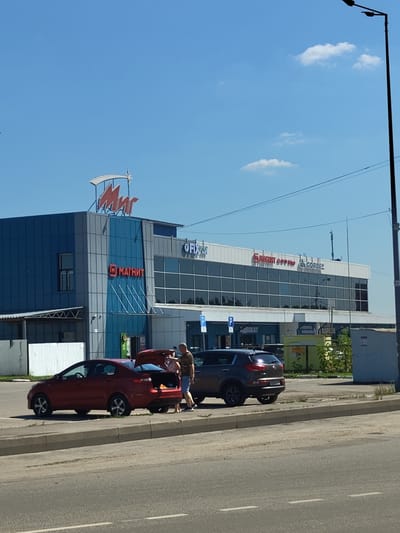 The image shows a sunny day in front of a retail complex in Zhukovsky, Russia. The complex has a large "Мир" (Mir) sign at the top, with the "Магнит" (Magnit) logo visible on the lower part of the building. Several cars are parked in front of the complex. A red sedan has its trunk open, and two people, a man and a woman, are standing near it. The man is wearing a camouflage t-shirt and shorts, while the woman is wearing a white top and shorts. They appear to be engaged in conversation or unloading items from the car. A dark gray SUV is parked next to the red sedan. The environment is urban, with a paved road and sidewalk in the foreground, and the retail complex and surrounding buildings in the background. A tall street lamp is visible on the right side of the image.