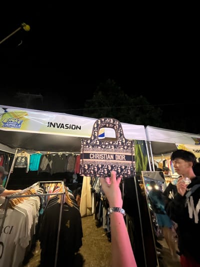 In Koronadal City, Philippines, an outdoor market stall is depicted at night. A person's arm, adorned with a watch, holds up a Christian Dior branded handbag in the foreground. Behind the handbag, a tent displays a banner with "INVASION" printed on it. Clothing racks with various garments are visible beneath the tent. In the background, a young man is seen holding what appears to be a small, clear object. The scene is illuminated by artificial lights, casting shadows and creating a nighttime ambiance. The ground is covered with what looks like grass or dirt.