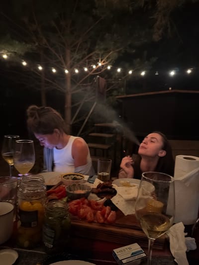A medium shot captures a dimly lit outdoor evening scene, likely in Taskino, Russia. Two women are seated at a table laden with food, drinks, and smoking paraphernalia. The woman on the left, with her hair pulled up, is looking down at her phone, illuminated by its screen. She is wearing a white tank top and a grey cardigan. In front of her are wine glasses and various food items, including what appears to be pickled vegetables in jars and sliced tomatoes. The woman on the right is exhaling smoke from a cigarette or similar device, her head tilted back and eyes closed. She is wearing a dark top. A roll of paper towels and a pack of HEETS are visible on the table. String lights hang in the background, casting a warm glow through the trees, suggesting a relaxed outdoor gathering.