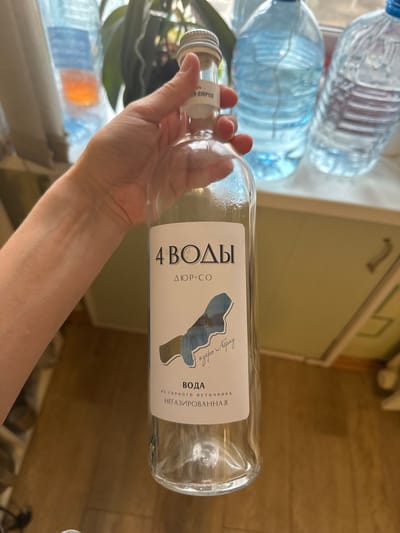 The image depicts a hand holding a glass bottle of "4 VODY" water, originating from a mountain spring near Lake Abrau. The bottle is labeled "ДЮР-СО" and indicates that the water is non-carbonated. The setting appears to be indoors, with other water bottles visible in the background. The city of Kladovitsy, Russia is not directly visible or identifiable in the image.