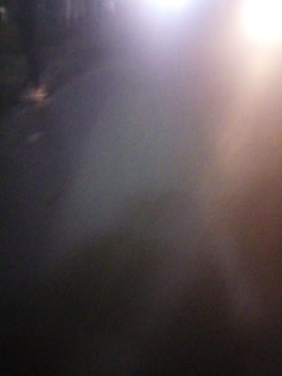 An indistinct image depicts an outdoor scene at night. Illumination from streetlights or similar sources creates a strong light flare in the upper portion, casting shadows across a paved surface, potentially a road or path. The heavy blur and low clarity render details unrecognizable, suggesting fog, rain, or a combination obscuring the environment. The composition consists mainly of gradients and blurred shapes, lacking sharp focus.