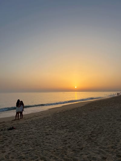 The image depicts a beach scene at sunset in Almancil, Portugal. The sky transitions from a light blue-grey at the top to vibrant orange and yellow near the horizon, where a bright, circular sun is positioned just above the water line. The ocean surface reflects the sunlight, creating a shimmering path of light across the calm water. Gentle waves break along the sandy shore, producing white foam.

In the left foreground, two female individuals are standing on the wet sand near the water's edge. Both appear to be wearing light-colored, short dresses or tunics. One individual has long dark hair, while the other has shorter or pulled-back dark hair. They are positioned close to each other, seemingly observing the ocean or interacting. On the drier sand slightly behind them, two dark-colored items, consistent with shoes or sandals, are present.

Further down the beach to the right, several indistinct figures are visible, some appearing to be in the water and others on the sand, indicating additional beachgoers in the distance. The beach itself consists of light-colored sand with visible textures and footprints.