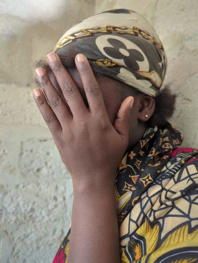 A close-up photograph depicts a person of African descent with dark skin and dark hair, partially obscured by a headscarf and hand. The individual is wearing a patterned headscarf featuring a repeating floral or clover-like design and a gold chain pattern, positioned over the left side of their head. The person's hand covers the lower half of their face. The ear is visible and adorned with a small gold stud earring. The person is wearing a colorful patterned garment with black, yellow, and red elements. The background is a textured, neutral-colored surface, possibly a wall. The location is Elele Alimini, Nigeria.
