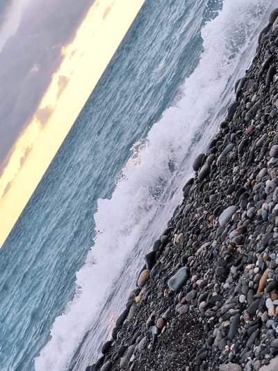 The image depicts a slightly tilted view of a pebble beach meeting the sea. The water is a muted blue-gray, with a breaking wave creating white foam near the shore. The beach is covered in numerous smooth, dark pebbles of varying sizes. In the upper left of the frame, the sky is a mix of grey clouds and a soft yellow band of light, suggesting either dawn or dusk. The location is identified as Zemo Kvariati, Georgia. There are no people or animals visible in the scene. The environment is a natural coastal landscape.