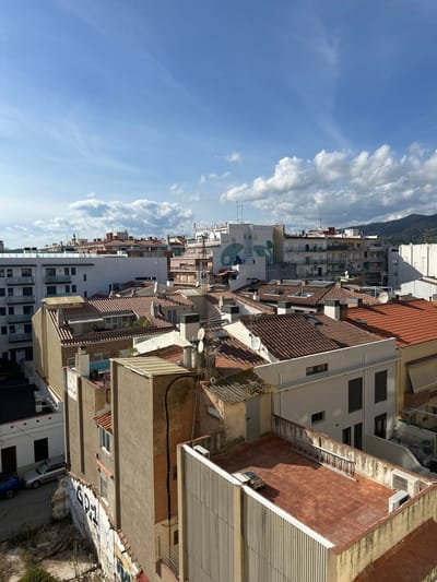 The image presents an elevated, wide-angle view of a densely constructed urban environment in Pineda de Mar, Spain, under a partially cloudy blue sky.

The scene is dominated by an array of buildings, primarily residential or mixed-use, with varying heights and architectural styles. A large proportion of the buildings feature pitched roofs covered in reddish-brown terracotta tiles. Numerous chimneys, ventilation shafts, television antennas, and satellite dishes are visible on the rooftops, including a prominent central antenna mast with multiple dishes.

In the immediate foreground, a flat, reddish-brown tiled rooftop is visible on the lower right, featuring an air conditioning unit, a white ladder leaning against a low wall, and a small metallic object. The adjacent building facade facing the viewer exhibits a light-colored, vertically ribbed texture. Below and to the left of this, an uneven plot of land with sparse vegetation is partially visible, alongside a retaining wall spray-painted with graffiti. A silver sedan and a blue vehicle are parked on an unseen street or alley in this lower left quadrant.

Moving into the mid-ground, a complex arrangement of buildings continues. Facades range in color from white to cream and light grey, with many featuring balconies and windows. One distinctive building in the mid-background displays a large, abstract mural in light green and light blue on its white exterior. A tall, narrow building in the mid-left features a dark, pipe-like conduit running vertically along its exterior.

The background shows the urban area extending into the distance, eventually meeting a horizon dominated by green, tree-covered hills or mountains. No people, discernible actions, or direct interactions are visible within the frame. The lighting suggests bright daylight conditions, casting shadows across the rooftops and buildings.