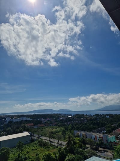The image shows a view from a high vantage point in Banyuwangi, Indonesia. The sky is predominantly blue with scattered white clouds. The sun is visible in the lower left corner. Below the sky, there is a strip of land containing buildings, vegetation, and a road. The structures vary in size and color, interspersed among green trees. A body of water lies adjacent to the land, separated by a thin strip of land in between.