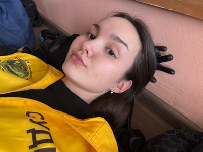 This image depicts a young adult female lying or reclining in an indoor setting in Tayshet, Russia. The individual has dark brown hair, fair skin, and light brown eyes, looking directly towards the camera. She is wearing a black long-sleeved garment beneath a bright yellow zippered vest. The vest features Cyrillic text, including "ФЕДЕРАЦИЯ СПОРТИВНОГО" and "ОБЛАСТИ" on the upper left, and "СУДЬЯ" on the lower right, along with a circular emblem showing a stylized creature with wings. A small hoop earring with a clear stone is visible in her left earlobe, and a smaller plain gold hoop or cuff is present higher on her left ear. Her head rests against a textured, light pink wall, which meets a dark brown wooden baseboard. A black gloved hand with fingers extended is visible near the top of her head against the pink wall. Various dark objects, including a blue bag and other straps, are present on the dark floor surface surrounding her.