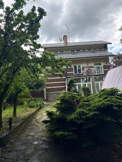 The image shows a residential area in Lesnoi Gorodok, Russia. In the foreground, there is a green bush and part of a gray object. Behind the bush, there's a multi-story building with a mix of brick and stone facade. The building has white-framed windows, a balcony with black railings, and exterior lighting fixtures. The roof is gray and has a chimney and a satellite dish. A TV antenna extends above the roof. Overhanging the building are several trees. A stone-paved path is visible on the left side of the image, and beyond it is a grassy area. The sky above is cloudy.