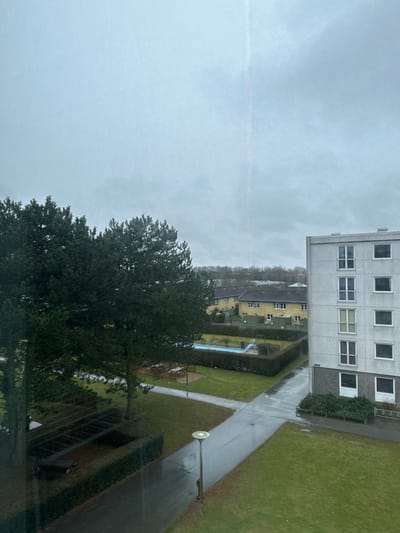 The image displays an outdoor residential area in Taastrup, Denmark, viewed from an elevated perspective under an overcast, light grey sky. The environment appears damp, with wet paved surfaces and patches of snow visible on grassy areas.
To the left, several tall coniferous trees stand, beneath which patches of snow are evident on the ground. Two distinct wooden picnic table sets are present: one partially obscured by trees on the far left, and another in a more open grassy section, both surrounded by low hedges. A single street lamp with a circular, frosted luminaire is positioned near a wet asphalt pathway.
On the right, a light grey, multi-story apartment building with a darker grey ground level features multiple white-framed windows, some with Juliet balconies. Directly across a paved path from this building, a rectangular swimming pool, filled with blue water and featuring a blue and white slide, is enclosed by a manicured hedge.
In the background, behind the pool and hedges, several yellow-sided residential houses with dark roofs are visible. The distant horizon is marked by a dense line of bare deciduous trees. No individuals are present in the scene.