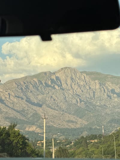 The image shows a wide shot of a mountainous landscape with a village nestled in the foothills. The mountains are rugged and rocky, with a hint of green vegetation on their lower slopes. The sky above is partly cloudy, with fluffy white clouds against a pale blue background. In the foreground, there are trees and power lines, suggesting a human presence in the area. The village appears to be composed of several houses with orange roofs, scattered among the trees. A telecommunications tower is visible on a hillside, further indicating modern infrastructure. The overall scene evokes a sense of remote, natural beauty mixed with signs of civilization.