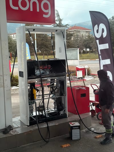 The image shows a gas station in Serinhisar, Turkey, with a worker repairing a gas pump. The pump is open, revealing its internal components and wiring. A black hose extends from the pump towards the worker. A container of Wunder oil is near the pump. The worker is wearing dark clothing with reflective stripes on the pants. The gas station has a "Long" sign and a flag with text. In the background, there are trees and buildings, including a building with a red roof.