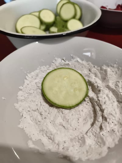 The image shows a close-up view of a white bowl filled with a light-colored powder, possibly flour or a breading mix, with a single slice of zucchini placed on top. The zucchini slice is round with a green rind and a pale green interior, showing the characteristic seeds and texture of the vegetable. In the background, another white bowl is visible, containing more sliced zucchini. The overall scene suggests food preparation, likely for frying or baking. The lighting is natural, indicating daytime, and there are no specific location cues beyond the general context of a kitchen or food preparation area. The mood is neutral, focusing on the ingredients and the early stages of cooking. No text is visible in the image.