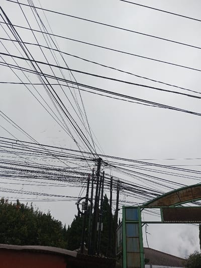 The scene, located in Ambakirano, Madagascar, captures an urban street view under an uniformly overcast sky. The upper two-thirds of the frame are dominated by a dense, tangled network of black electrical and communication wires, crisscrossing horizontally and diagonally. These wires converge upon and extend from a prominent, dark-colored utility pole positioned slightly left of the image center, which supports multiple smaller conduits and various attachment points. Below the utility pole and to its left, dense dark green trees and foliage are visible. In the lower left quadrant, a segment of a reddish-brown building facade or roofline is present. To the lower right, a partially visible green metal archway structure with a curved, rusted upper section and opaque lower panels extends into the frame from the right edge. Behind this archway, a section of a light-colored tiled roof is visible. No individuals or direct actions are depicted.