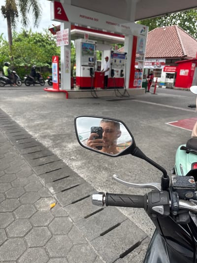 A person's face is reflected in the side mirror of a motorcycle, holding a smartphone and seemingly taking a picture. The location is recognizable as a Pertamina gas station, with gas pumps and the station's building in the background. Several other motorbikes are parked nearby. The pavement where the motorcycle is standing is made of interlocking hexagonal stones, indicating a common type of road surface in Munggu, Indonesia.