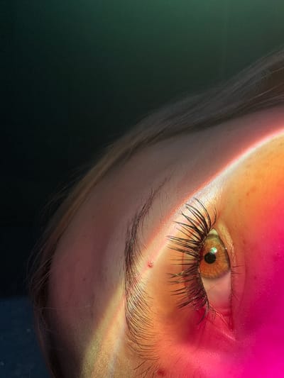 An extreme close-up shot focuses on the right eye of a person, with a portion of their forehead and eyebrow visible. The iris of the eye is a warm hazel color, and a small, iridescent speck is reflected in the pupil. Long, dark eyelashes frame the eye. A diagonal band of colored light, transitioning from yellow to pink, illuminates the skin on the forehead and cheek. The background is a dark, mottled green. No location in Velykyi Sukhodil, Ukraine is discernible from the image.