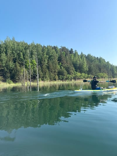 A man stands on a yellow and white GO BOARD paddleboard on a body of water, possibly a lake or wide river, adjacent to a forested area under a clear blue sky. He is wearing a black shirt, blue shorts, and a black hat. He is holding a two-bladed paddle over his head with one hand. The forest is densely packed with green trees and the water reflects the trees and sky. The photo is likely taken at a low angle, close to the water's surface. The location in the city of Dzerzhinsky, Russia is not discernible from the image.