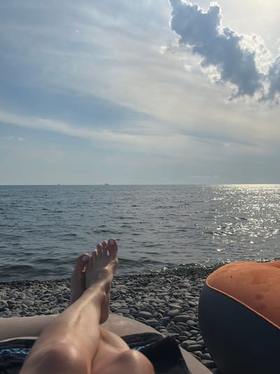 A close-up shot captures a pair of bare feet with white-painted toenails resting on a pebble beach. The feet are positioned in the foreground, with legs extending out of frame. Behind the beach, a calm body of water stretches to the horizon, reflecting the sunlight in a shimmering path. Two cargo ships are visible in the distance on the water. The sky above is a mix of blue and white clouds, with the sun casting a bright glare. The scene appears to be set in Shirokaya Balka, Russia, suggesting a peaceful day by the sea. An inflatable orange and black object is partially visible on the right side of the frame.