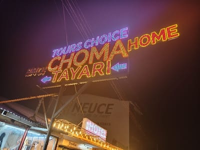 A vibrant, multi-colored neon sign reading "TOURS CHOICE CHOMA HOME" and "ENJOY TAYARI" is prominently displayed. Arrows indicate directions, and thin wires extend from the sign's structure. Below, a hotel sign with glowing lights illuminates a building. A large sign in the background bears the word "NEUCE". The overall scene is set against a dark sky, suggesting nighttime.