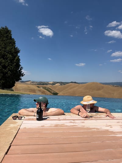 Two individuals are relaxing in an infinity pool overlooking a rolling landscape of dry, golden hills under a clear blue sky with scattered white clouds. The person on the left wears a green baseball cap and leans on the pool's edge, with a bottle of wine and a glass next to them. The person on the right wears a straw hat and holds a glass of champagne. The pool has a wooden deck edge, and sunglasses are placed on the left side of the deck. The environment suggests a warm, sunny day in the Tuscan countryside near Asciano, Italy, known for its distinctive clay hills known as the Crete Senesi.
