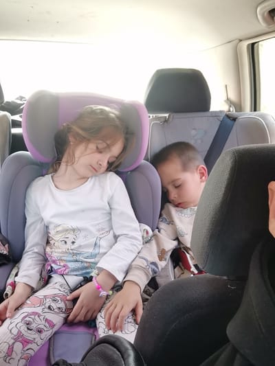 Two children, a girl and a boy, are sleeping in the back of a vehicle. The girl is in a purple and grey car seat and is wearing a white long-sleeved shirt with an Elsa Frozen graphic and Paw Patrol patterned pants. She has a pink and purple beaded bracelet on her left wrist. The boy is also in a grey car seat, wearing a long-sleeved pajama top with a dinosaur pattern. The scene appears to be inside a car during daylight. The location is not determinable from the image, and no evidence suggests the city of Tsarkva, Bulgaria.
