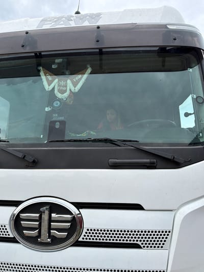 The image displays the front and upper side of a large, white FAW truck, observed from an elevated viewpoint. The truck is currently stationary, with a clear, bright sky in the background suggesting daytime.
A prominent feature is the truck's chrome oval emblem on the front grille, which contains a stylized, layered character, characteristic of the FAW brand logo. The grille itself consists of vertical white slats with black, honeycomb-perforated sections.
Through the expansive windshield, the interior of the truck's cab is partially visible. A young child's face is faintly discernible, looking out from inside. Hanging from the top of the windshield is a decorative, fringed pennant, bearing the text "FAW" repeated several times, along with a logo similar to the main FAW emblem. Other items visible on the dashboard include a phone holder and what appears to be a dash camera or similar electronic device mounted near the top of the windshield. A steering wheel can also be faintly seen within the cab. Two black windshield wipers are positioned at the base of the windshield.
On the upper side panel of the truck, above the windshield, the word "FAW" is painted in large, grey lettering. The overall scene suggests the truck is parked outdoors in Kamyennoye, Russia, under good weather conditions.
