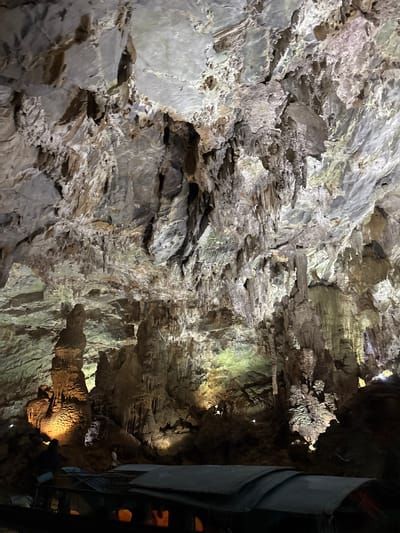 The image shows the interior of a cave in Phong Nha Commune, Vietnam, with a boat in the foreground. The cave is characterized by its large stalactites and stalagmites, illuminated by artificial lighting that highlights the rock formations in earthy tones of brown, gray, and white, with patches of green and yellow. The ceiling is heavily adorned with numerous stalactites of varying lengths and thicknesses. Prominent stalagmite formations rise from the cave floor, some appearing massive and columnar.

In the foreground, the dark-colored roof of a boat stretches across the bottom portion of the image, partially obscuring the cave floor. Several orange life vests are visible through openings in the boat's covering, suggesting passengers are present. Two individuals are dimly visible standing on or near the boat. The lighting creates a dramatic contrast between the brightly lit formations and the shadowy depths of the cave. The overall impression is one of a vast, ancient natural wonder being explored by human visitors.