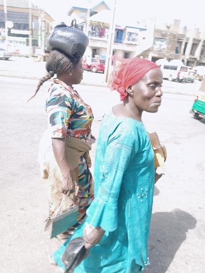 Two adult females are depicted walking on a paved surface in Mumbwa, Zambia, under bright daylight. The woman on the left, facing right, wears a multi-colored patterned shirt, a light-colored wrap around her waist, and dark hair styled with a high bun and a long braid. She carries a black plastic bag balanced on her head and a silver-toned handbag with a chain strap in her right hand. The woman on the right, also facing right, wears a red headscarf and a long-sleeved, turquoise-colored dress with patterned details and white bead embellishments. She holds a crumpled black plastic bag in her left hand and has a tan-colored item over her right shoulder. The background features multi-story buildings, consistent with an urban environment, and several vehicles including a red pickup truck, a white van, and a green and yellow tuk-tuk. Strong shadows indicate a bright, sunny day.