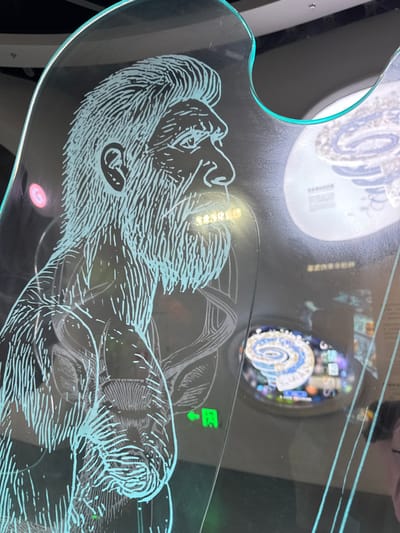 The scene captures an interior view of an exhibit display within a facility, likely a museum or science center, located in Guangming District, China. The central subject is a large, irregularly shaped transparent panel, possibly acrylic or glass, featuring a light blue-green etched outline of a man's head and upper torso in profile, facing right. The figure is depicted with a prominent beard, mustache, and hair, characteristic of an early human or hominid. Below the head, etched lines suggest internal anatomical structures, potentially a DNA helix or vascular system, and a partial outline of an animal, possibly a deer with antlers, is visible further down the panel. Small, yellow-orange illuminated text spelling "SOSORG" is present near the figure's mouth area. A small green illuminated icon, depicting an arrow pointing left alongside a character, is visible on the panel to the right of the figure's torso. The background, visible through and reflected on the panel, contains other illuminated displays. One prominent circular display features a detailed, multi-colored spiral graphic, resembling a brain or complex biological system, accompanied by Chinese text below it. The environment is indoors, with controlled lighting highlighting the exhibits and producing reflections on the panel's surface. A faint, blurred reflection of a person's head is visible in the lower right corner of the panel.