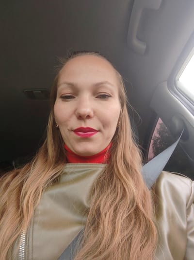 A woman is captured in a selfie taken inside a car, likely in Chukhlem, Russia. She is seated in what appears to be the front passenger or driver's seat, with a dark grey seatbelt diagonally crossing her chest. The woman has long, wavy hair of a light brown or blonde hue, which cascades over her shoulders. She is wearing a vibrant red turtleneck top beneath a light brown or tan jacket, possibly made of leather or a similar material. Her lips are adorned with bright red lipstick, and she wears small, gold-colored hoop earrings. Her expression is neutral to slightly pleasant, with a subtle hint of a smile as she looks directly at the camera.
The car's interior serves as the background, featuring a grey headliner. On the right side, an overhead grab handle is visible, and on the left, a portion of an overhead console or rearview mirror assembly can be seen. Light from outside the car suggests it is daytime, with a glimpse of blurry foliage or trees visible through the car's tinted window on the right. No discernible text is visible within the image.