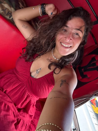 A woman with dark, curly hair and a bright smile poses for a selfie from inside a red auto-rickshaw in Kampot, Cambodia. She is wearing a red dress and several pieces of jewelry, including a beaded necklace and bracelets. She has tattoos on her chest, arm, and forearm. The auto-rickshaw's interior is red, with some camouflage fabric visible on the seating. The scene suggests a casual, outdoor setting, likely a street in Kampot, given the presence of the auto-rickshaw, a common mode of transport in the region.