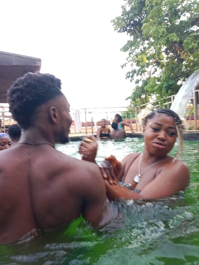 A group of people are enjoying a swimming pool with green water. A man with a high-top fade hairstyle and a well-defined back stands in the foreground facing left. A woman with blonde-streaked braids and a silver necklace looks at the camera while in the water. Other people are seen in the background, some near a waterfall feature. The pool is surrounded by a railing, and trees are visible in the upper right corner.