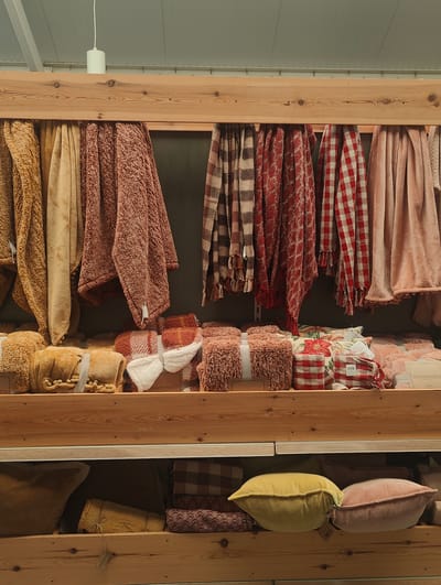 The image captures a scene inside a shop in Colchester, United Kingdom. The focus is on a wooden shelving unit filled with textiles, including throws and cushions. The top shelf showcases hanging throws in various colors and patterns, such as brown, beige, pink, plaid, and red gingham. Below the hanging throws are neatly stacked throws and cushions of similar colors and patterns, some adorned with tags and tied with ribbons or twine. The bottom shelf displays an assortment of cushions, including yellow and pink velvet pillows, along with other patterned textiles. The overall impression is that of a home goods or lifestyle store, specializing in soft furnishings. A white light fixture is visible on the ceiling above the shelves.