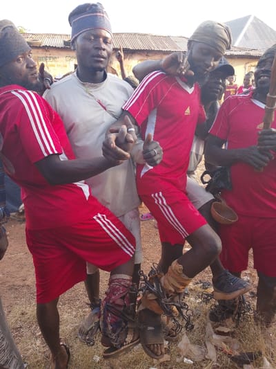 The image depicts an outdoor scene in Vodni, Nigeria, featuring a group of approximately eight adult males. Three individuals are prominent in the foreground. The central figure wears a light-colored tunic, a striped knitted cap, and a thin necklace, holding the hands of the two men flanking him. The man to his left wears a red Adidas t-shirt and shorts with white stripes, a brown fabric headwrap, and a silver-colored ring on his left ring finger; his left hand clasps the central figure's hand, while his right thumb is raised. His left ankle is visibly bound with chains wrapped in fabric over a sandal. The man to the central figure's right also wears a red Adidas t-shirt and shorts with white stripes, a brown fabric headwrap, and a silver-colored ring on his left ring finger; his right hand clasps the central figure's hand, and his right leg is similarly bound with chains wrapped in fabric, a black sneaker visible on his foot. This man smiles broadly. Another male figure to the far right, partially obscured and also in a red shirt, holds a wooden staff and has a small calabash gourd attached to his waist.

The ground is dry, reddish-brown dirt with sparse patches of dry grass and scattered white plastic debris. In the background, a weathered building with a corrugated metal roof is visible, bearing a partially legible sign that begins with "BIDEF". The overall lighting suggests a bright, sunny day. The men's expressions range from neutral to smiling.