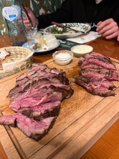 Two portions of sliced, rare grilled steak rest on a wooden cutting board. Small dishes containing white condiments, possibly salt and sauce, sit beside plates of salad and butter. A partially filled water bottle and a woven basket with bread are also visible. A person's hand is seen near the dishes.
